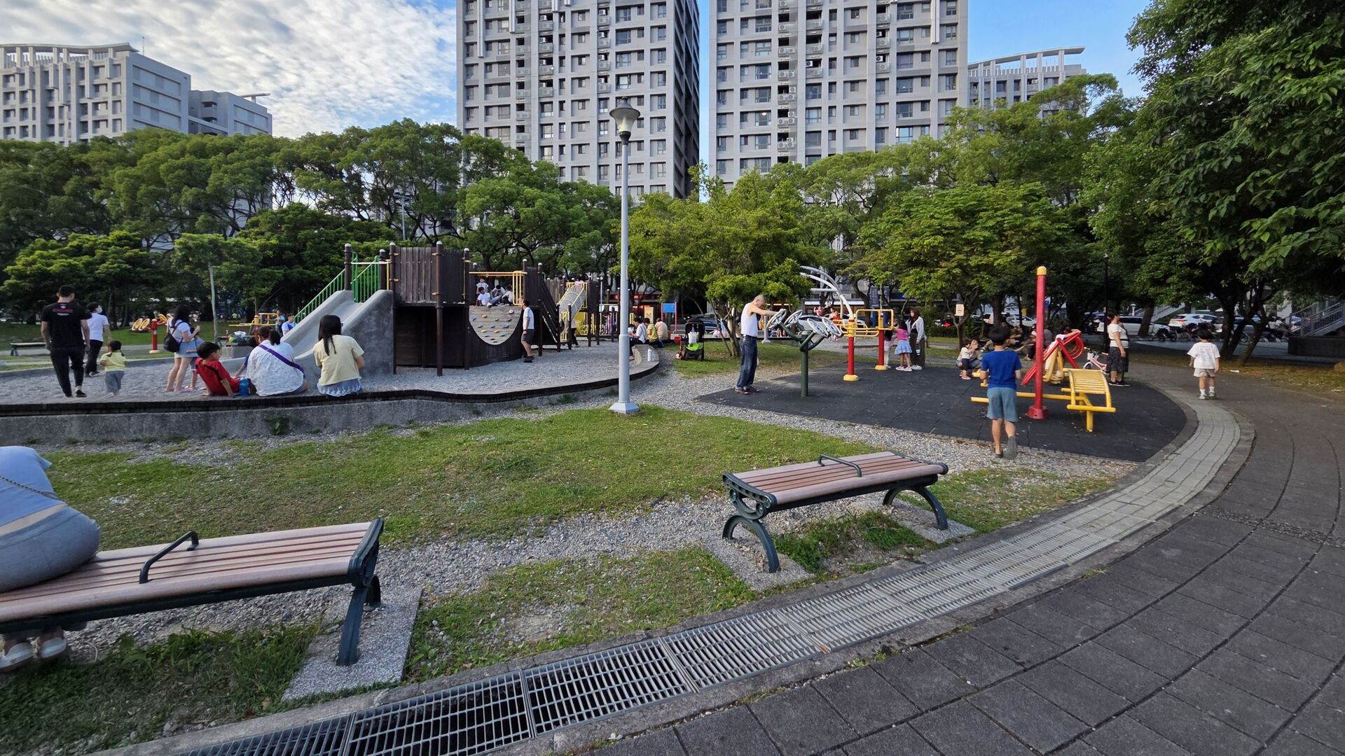 一個座落於高樓住宅區旁的社區公園，許多民眾正帶著孩子在綠地間的兒童遊樂區與健體設施周邊休閒活動。