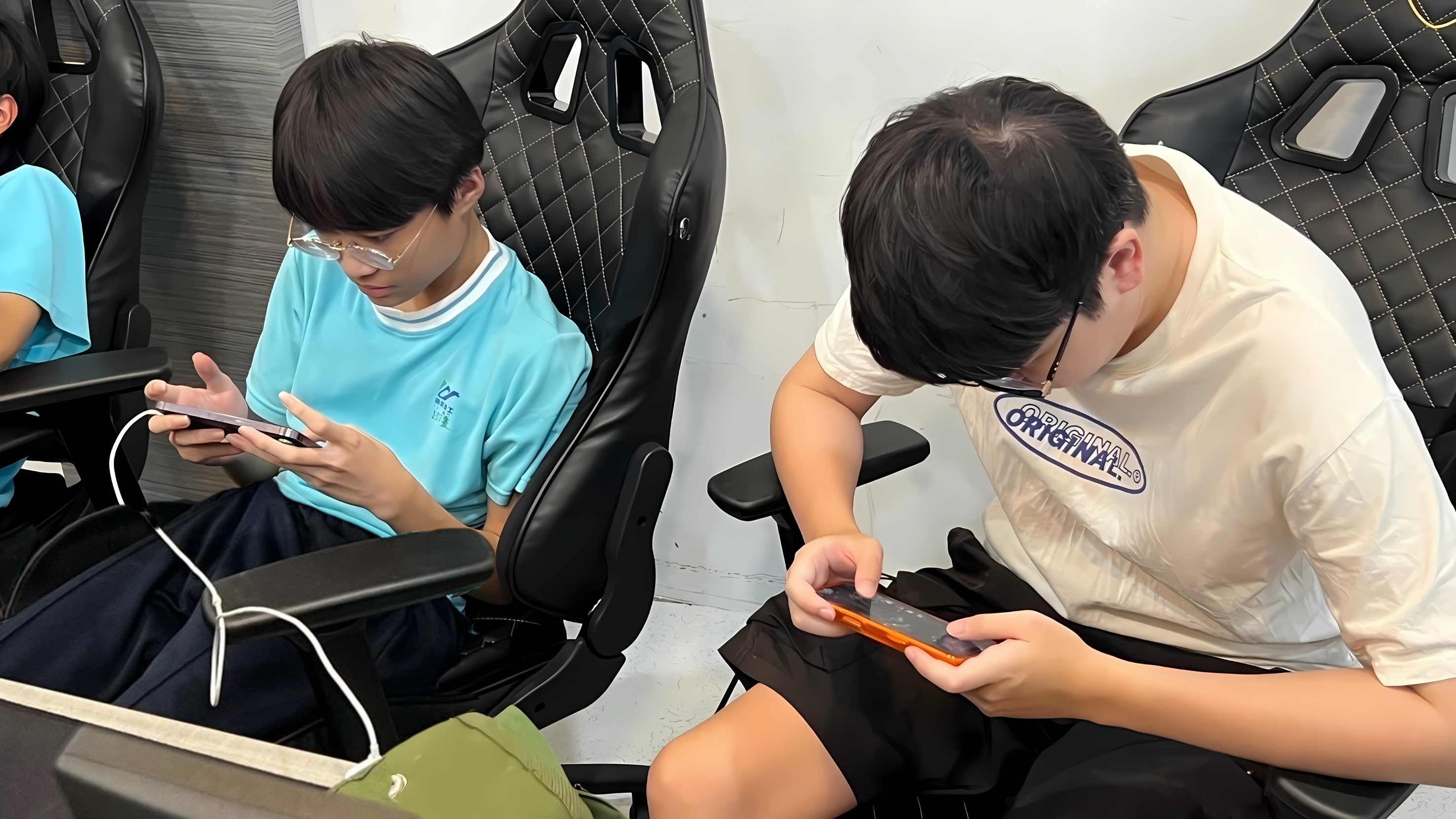 兩位學生正坐在黑底菱格紋電競椅上專注地操作著手中的智慧型手機，進行手機遊戲的對戰。
