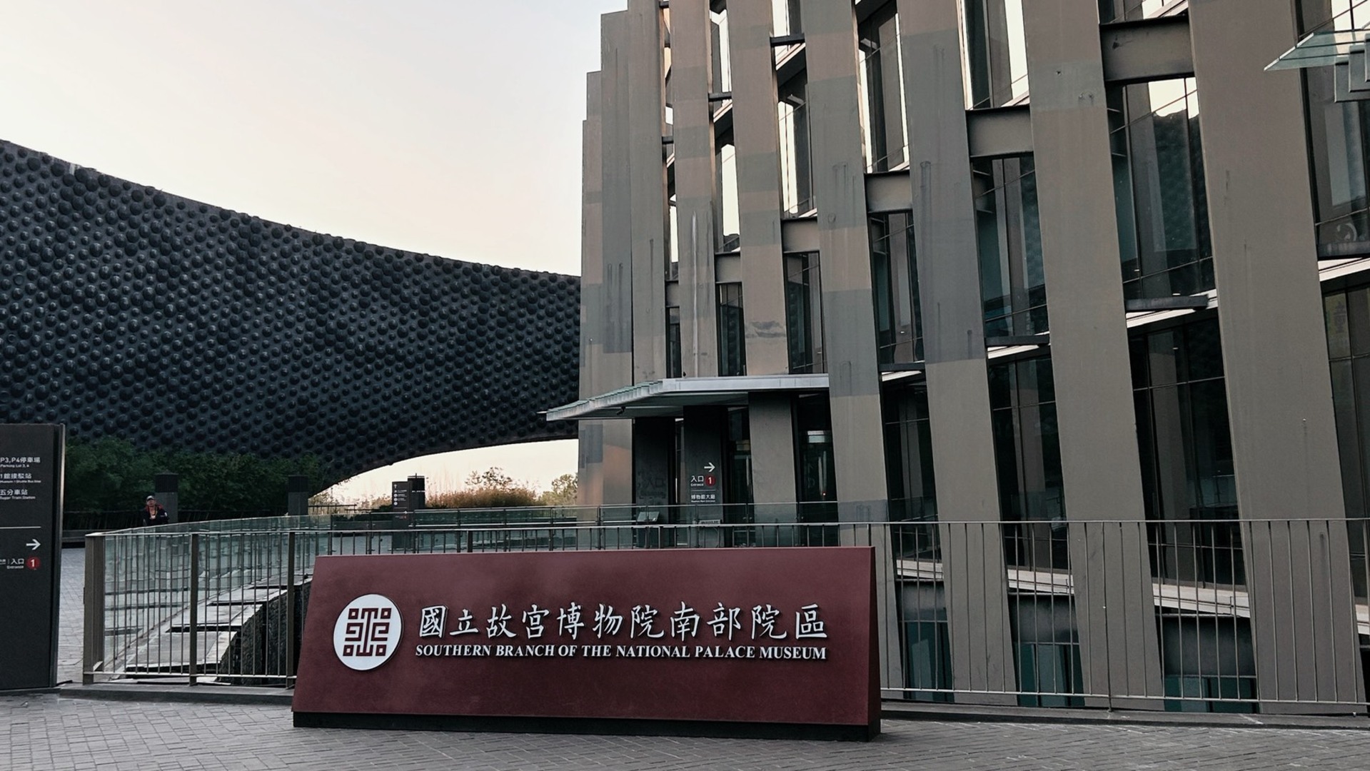 國立故宮博物院南部院區，前方立著紅褐色的院名招牌，後方是具現代設計感的灰色流線型建築與玻璃幕牆。