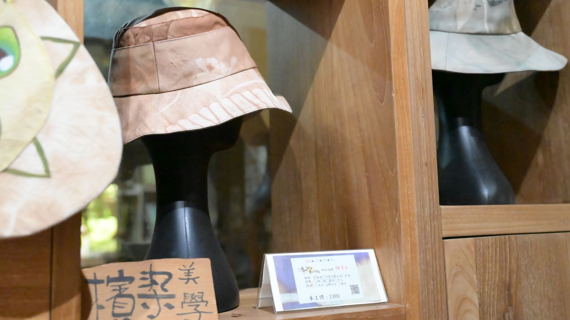 一個木製展示櫃中,陳列著一頂帶有植物葉片染印圖案的淺大地色漁夫帽,帽子戴在一個黑色的假人頭像上。