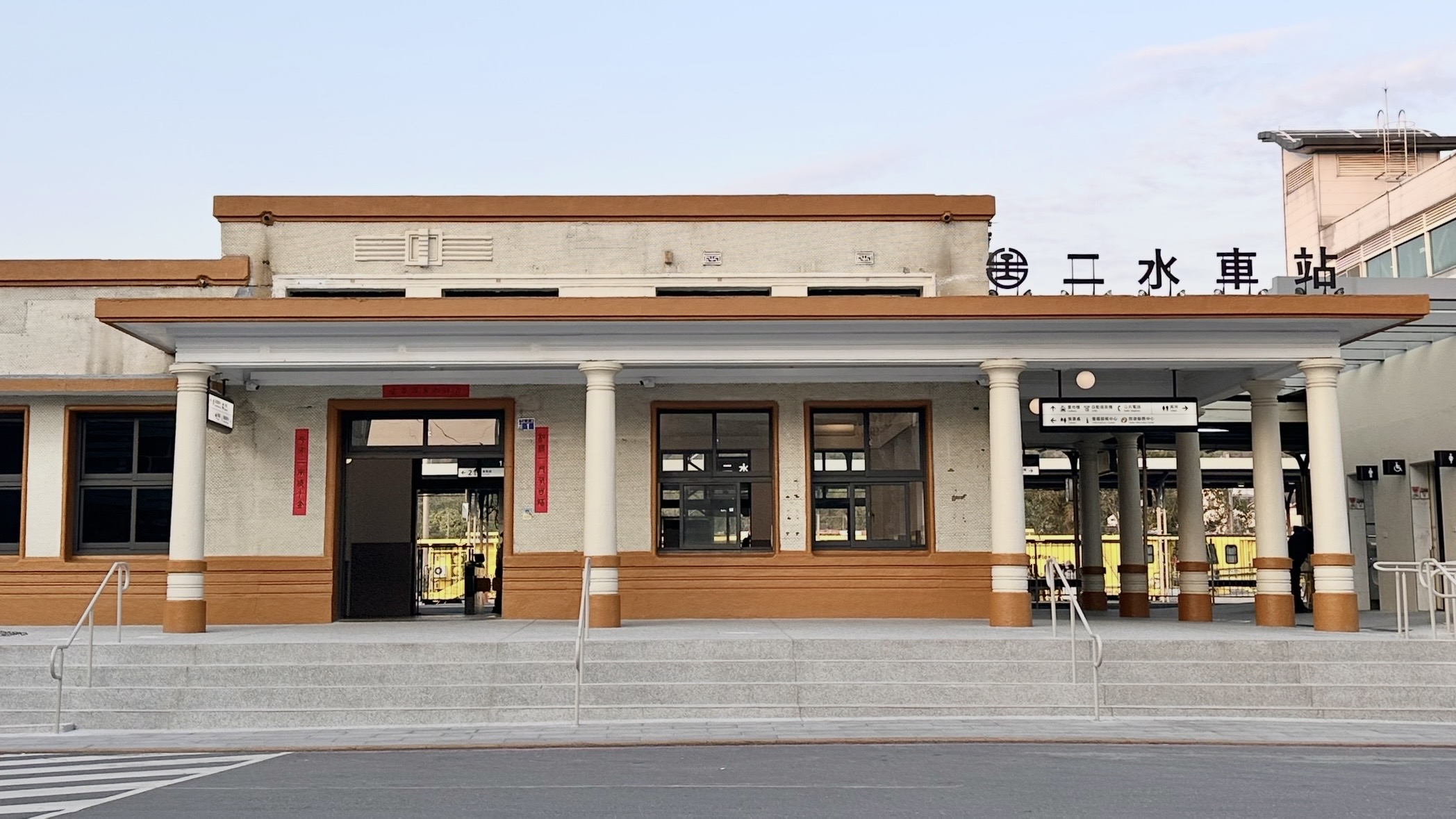 從另一角度看二水車站，呈現平整的灰牆與橘色邊框建築，門口貼有紅聯，右側上方可見台鐵標誌與「二水車站」字樣，整體環境整潔且明亮。