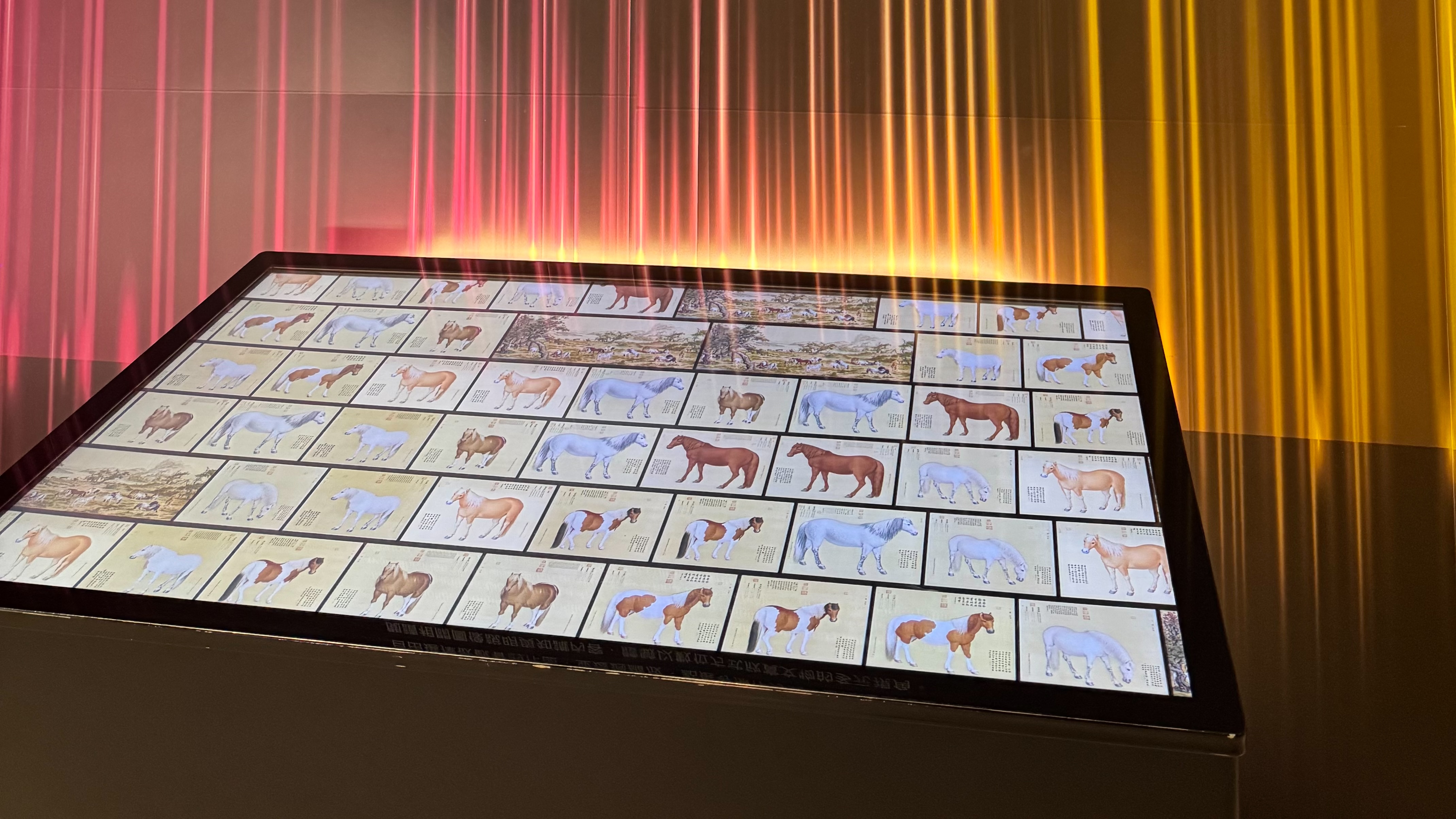 展區內的觸控螢幕上呈現許多格馬匹畫作，背景有紅黃色調的線條燈光效果。