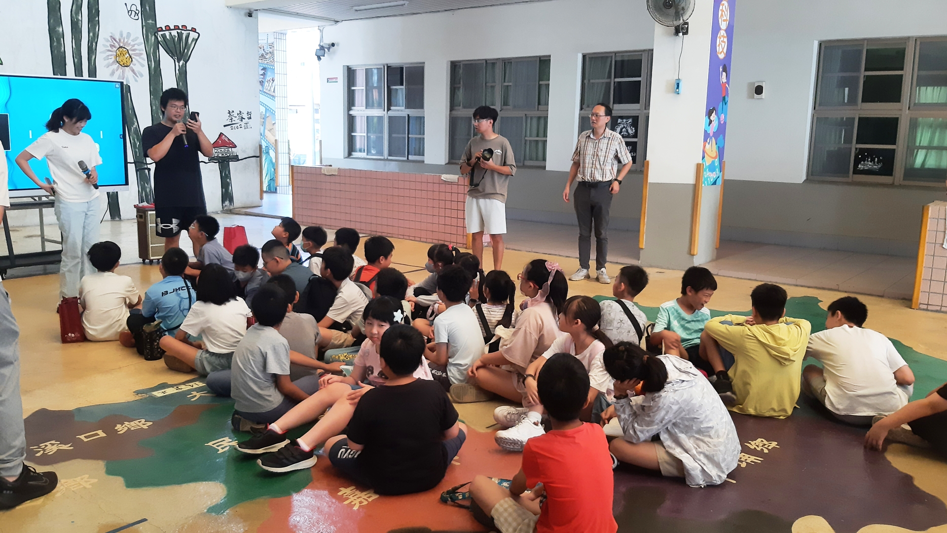 小朋友們坐在地上，前方站在電視螢幕前的大哥哥、大姐姐們拿著麥克風講話