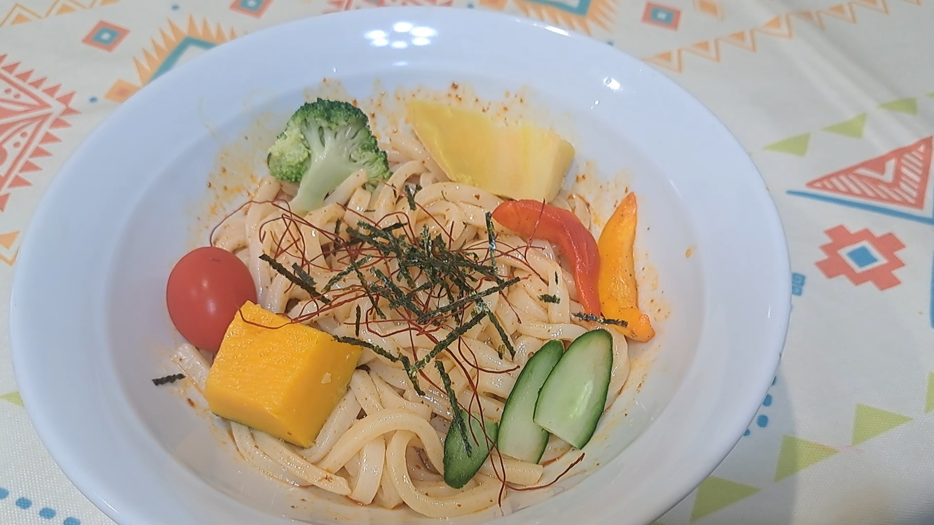 白色大碗中裝有烏龍麵，上面鋪著各式色彩鮮豔的蔬菜配料，包含花椰菜、南瓜塊、小蕃茄、小黃瓜片、彩椒，並撒上細海苔絲與辣椒絲裝飾。