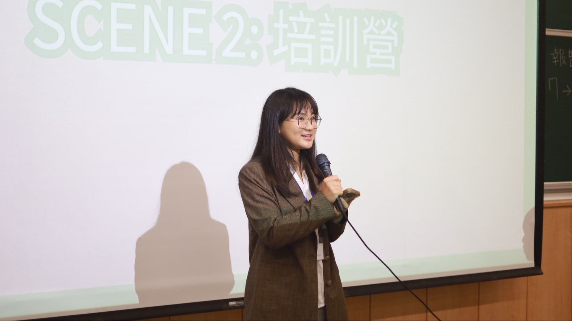 張育綺站在投影目前拿著麥克風演講