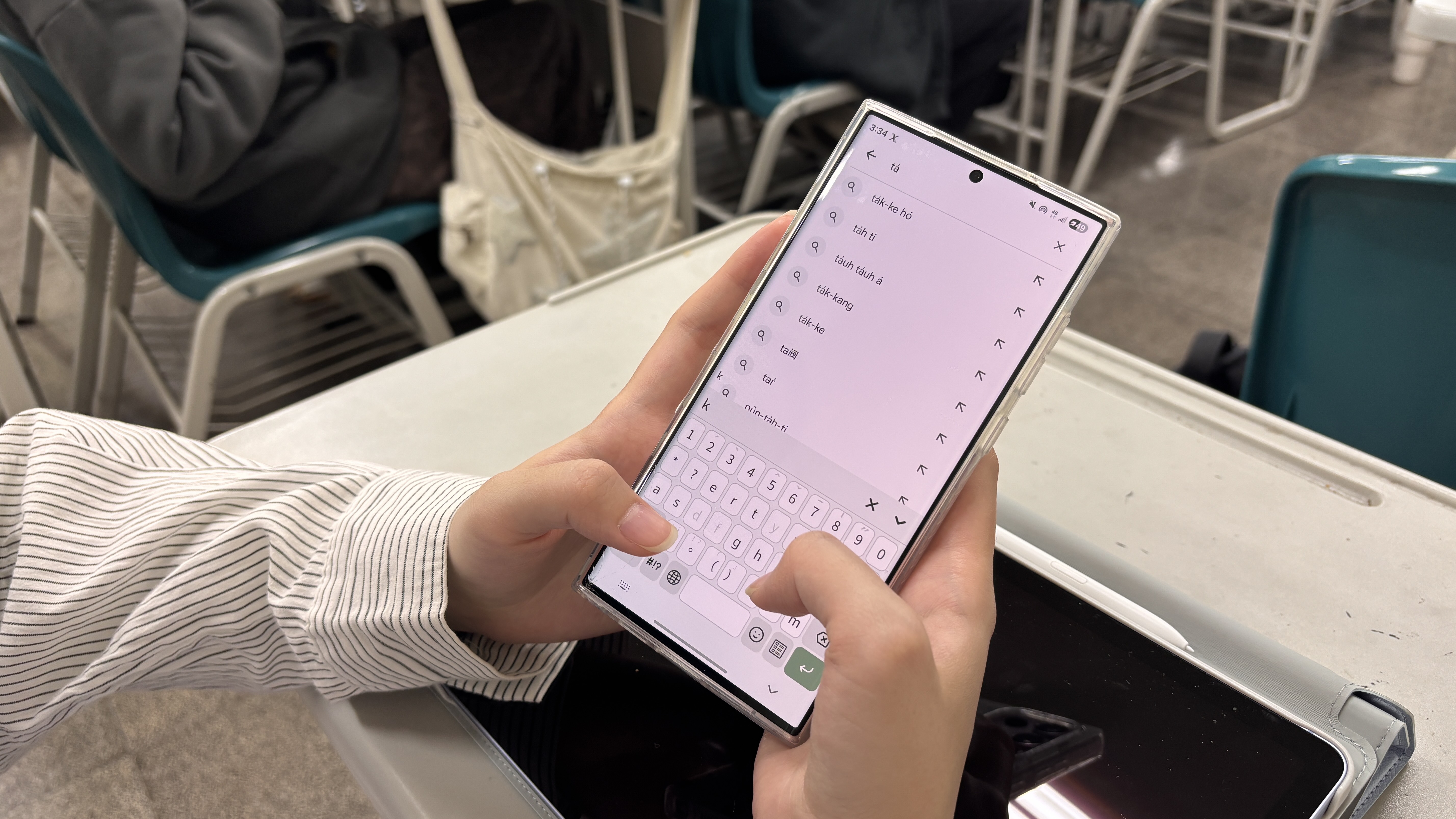 在教室課桌前,一人雙手操作手機,正使用羅馬拼音輸入法進行搜尋。