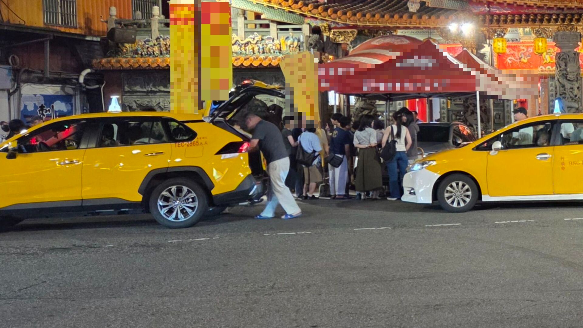 夜晚的廟宇前，一名男子正站在黃色計程車後方整理後車廂，背景有聚集的人群與紅色的活動帳篷。