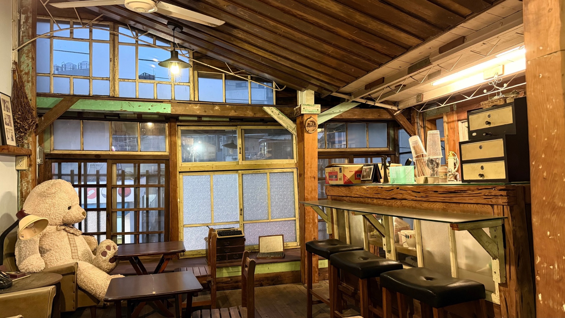 這是一個充滿復古木質調的室內空間，左側座位區放著一隻巨大的淺棕色大熊玩偶，右側則是木造吧檯與高腳椅，搭配老式窗框與溫暖的黃光吊燈，營造出悠閒的懷舊氛圍。