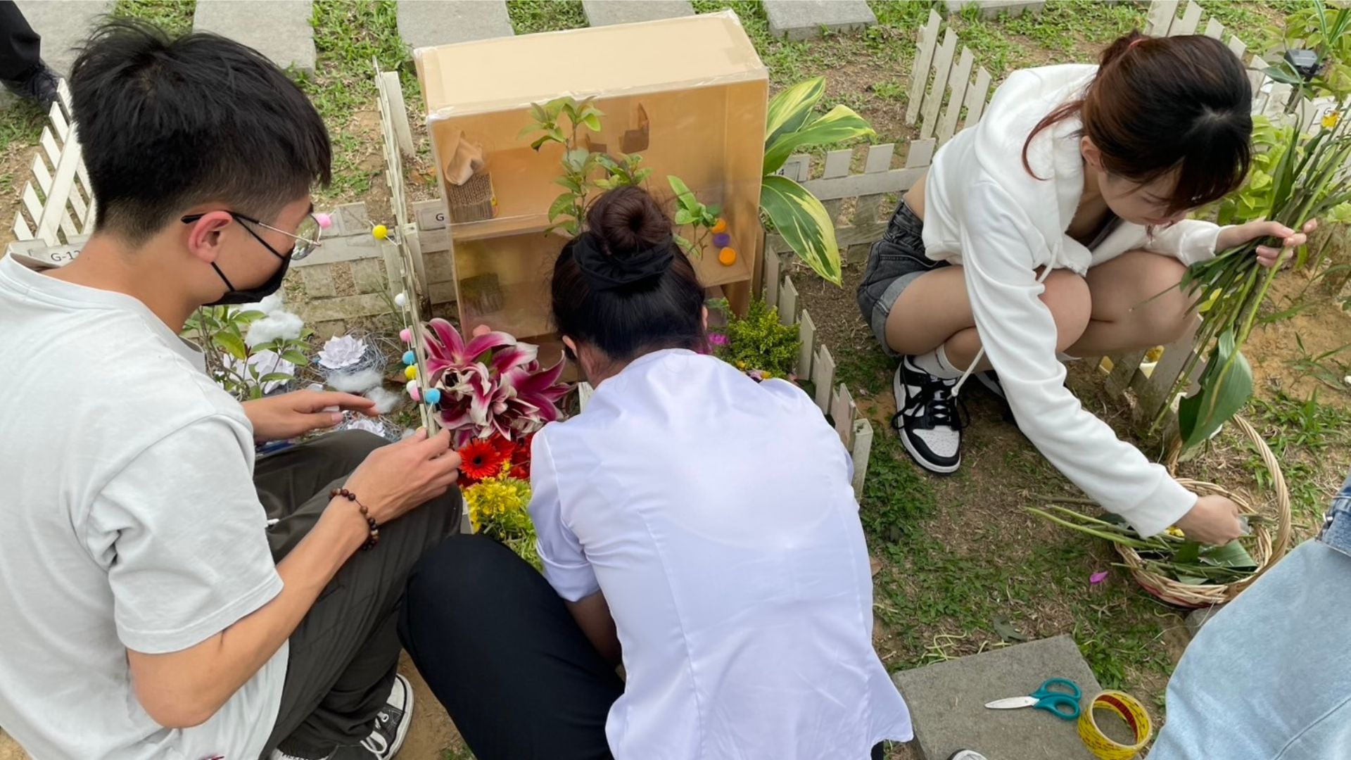 寵物殯葬與祭奠的側拍照片。畫面中有三位年輕人正蹲在草地上的寵物墓位前，細心地佈置與整理。