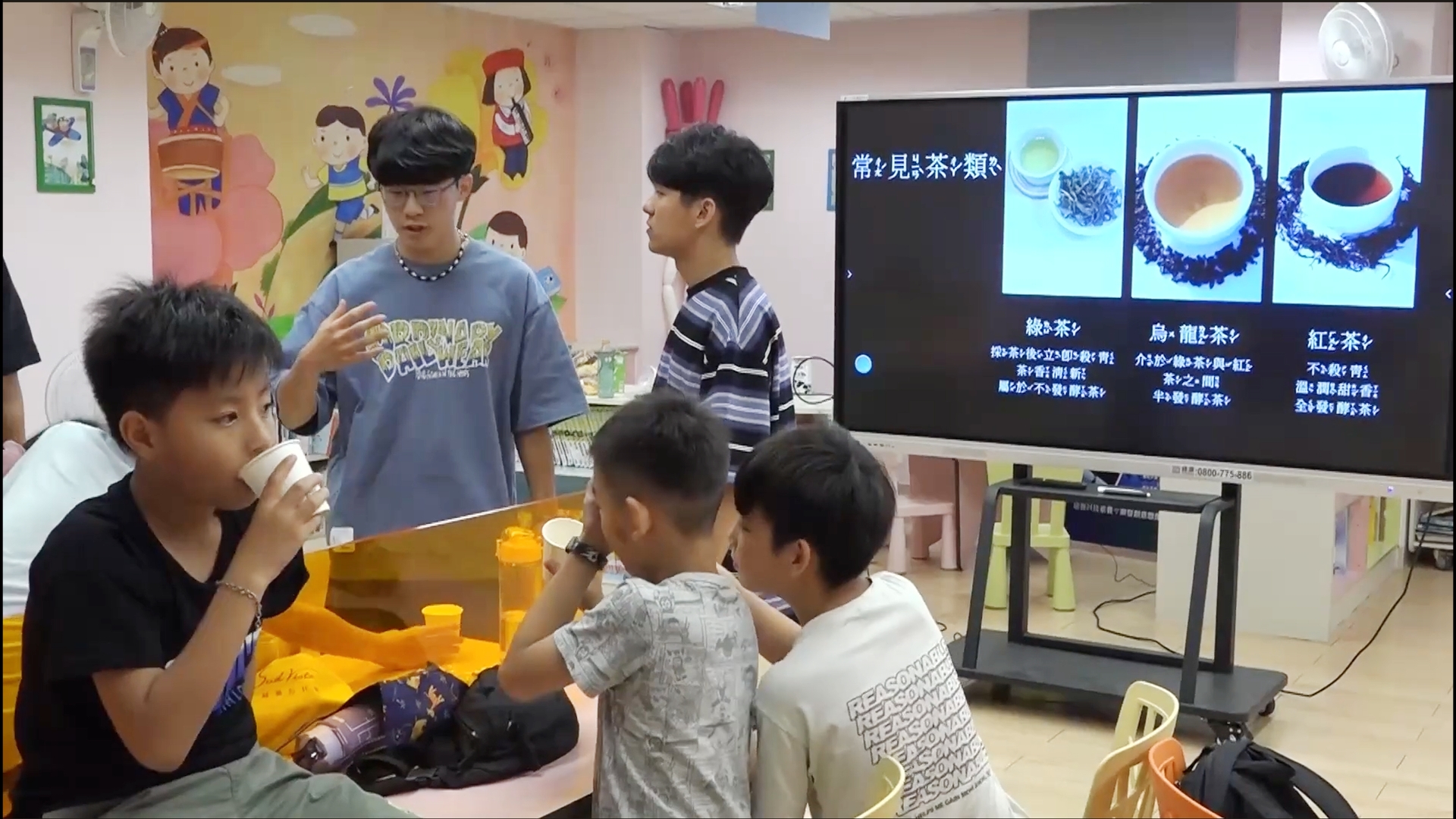 前方的電視上有綠茶、烏龍茶與紅茶三種常見茶類，兩位大學生正在引導孩子們試喝