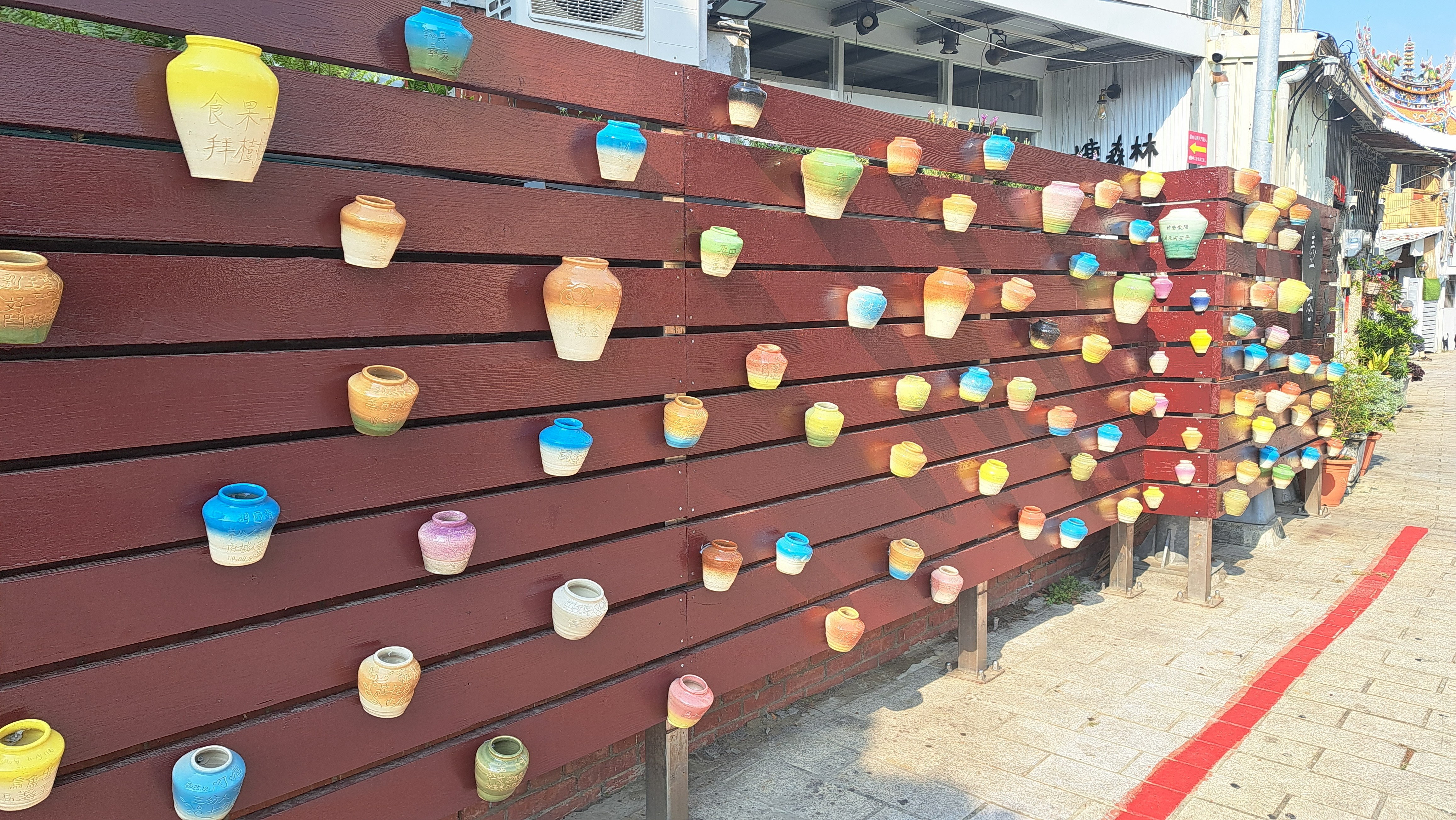 一面充滿藝術感的紅褐色木柵欄牆，牆上錯落有致地裝飾著許多五彩繽紛的小型陶罐。