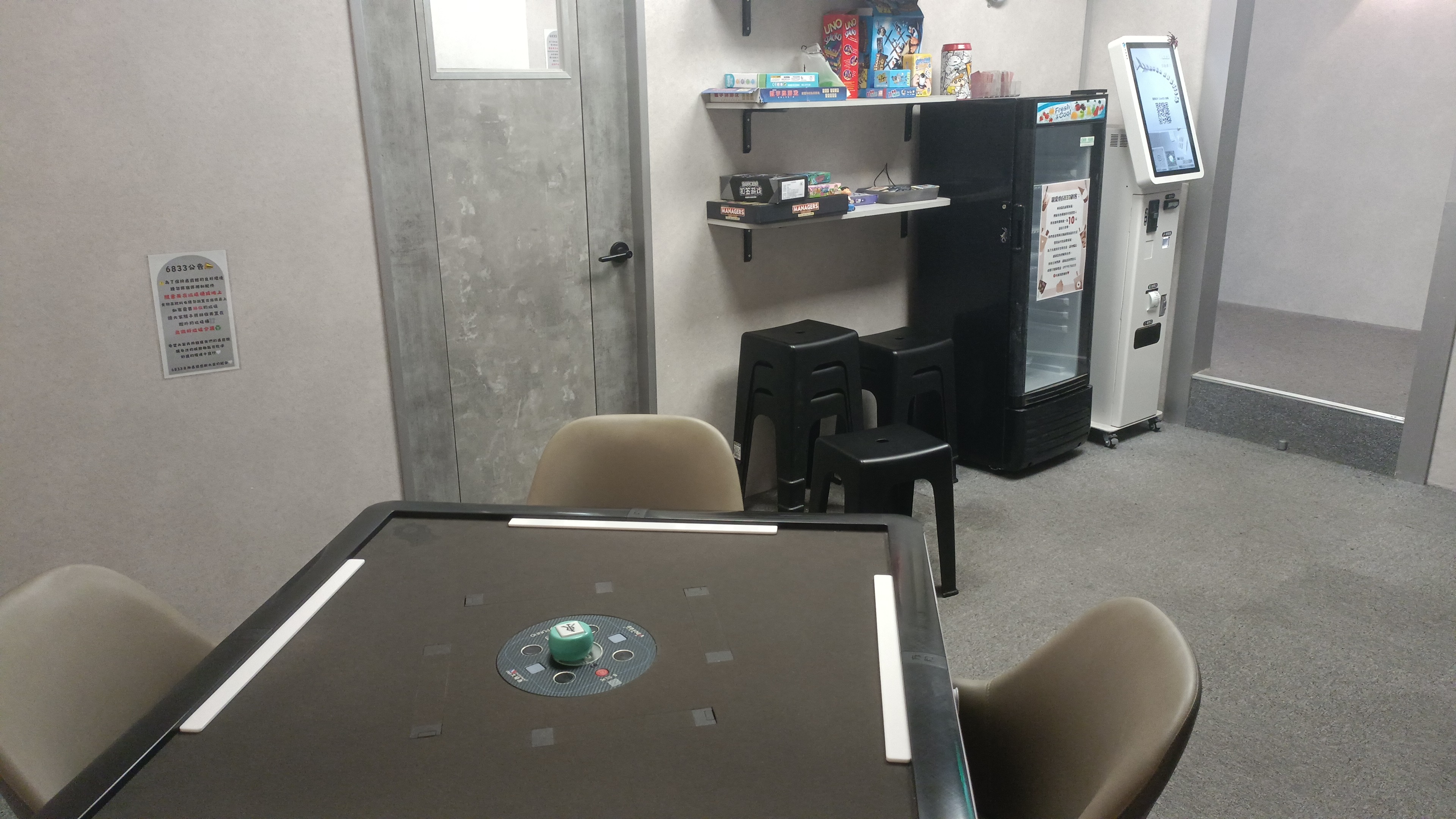 一個室內遊憩空間，前方有一張自動麻將桌，後方則設有放置桌遊的層架、小冰箱及一台白色自助機台。