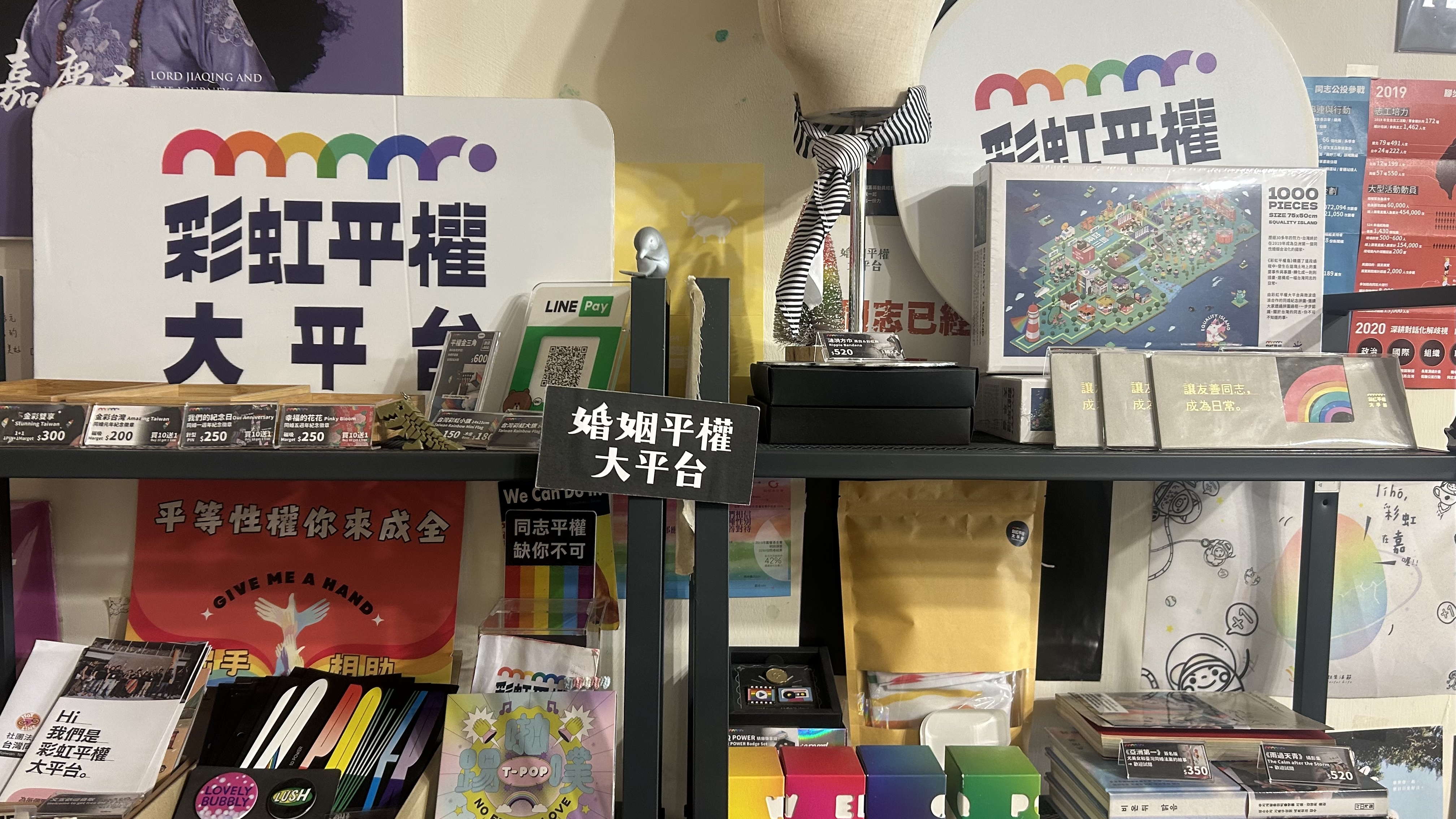 「彩虹平權大平台」辦公室或義賣活動現場的層架，上面擺滿了各式同志平權運動的宣傳品、文宣物資與紀念商品。