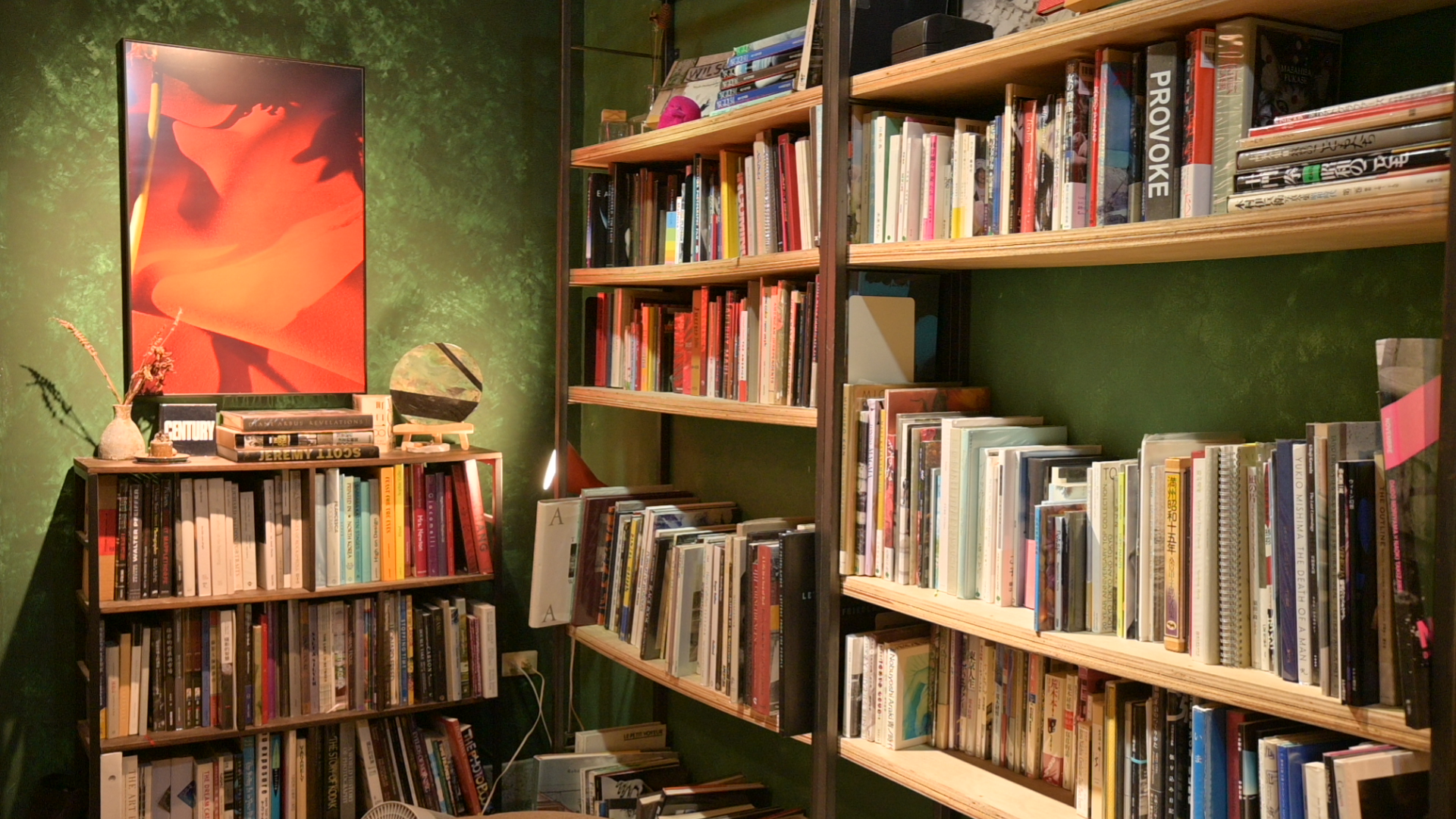 室內一隅設有幾座裝滿書籍的木質書架,牆面漆成深綠色,左側掛著一幅橘紅色調、光影強烈的藝術攝影作品。