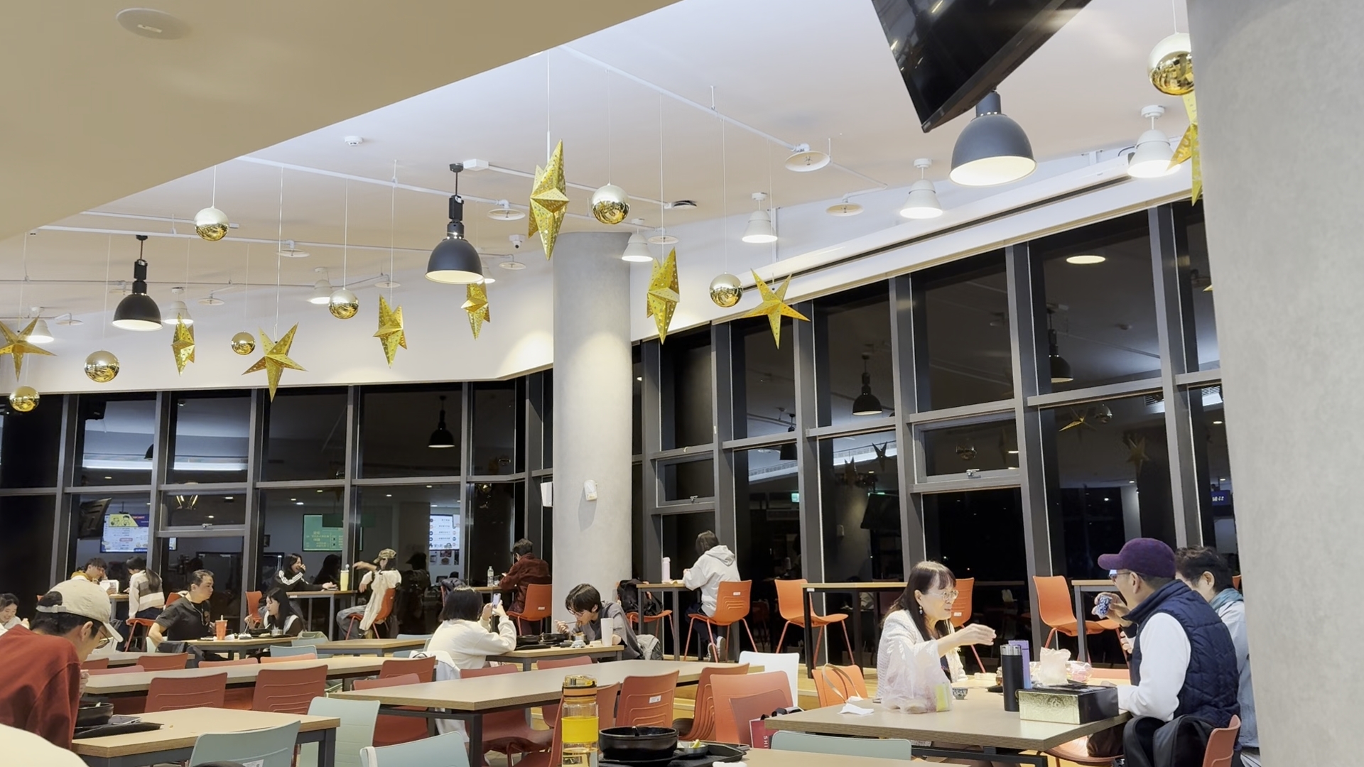 公共用餐空間內有許多學生或民眾分坐在餐桌前用餐，室內挑高寬敞，天花板垂掛金色星星與燈飾