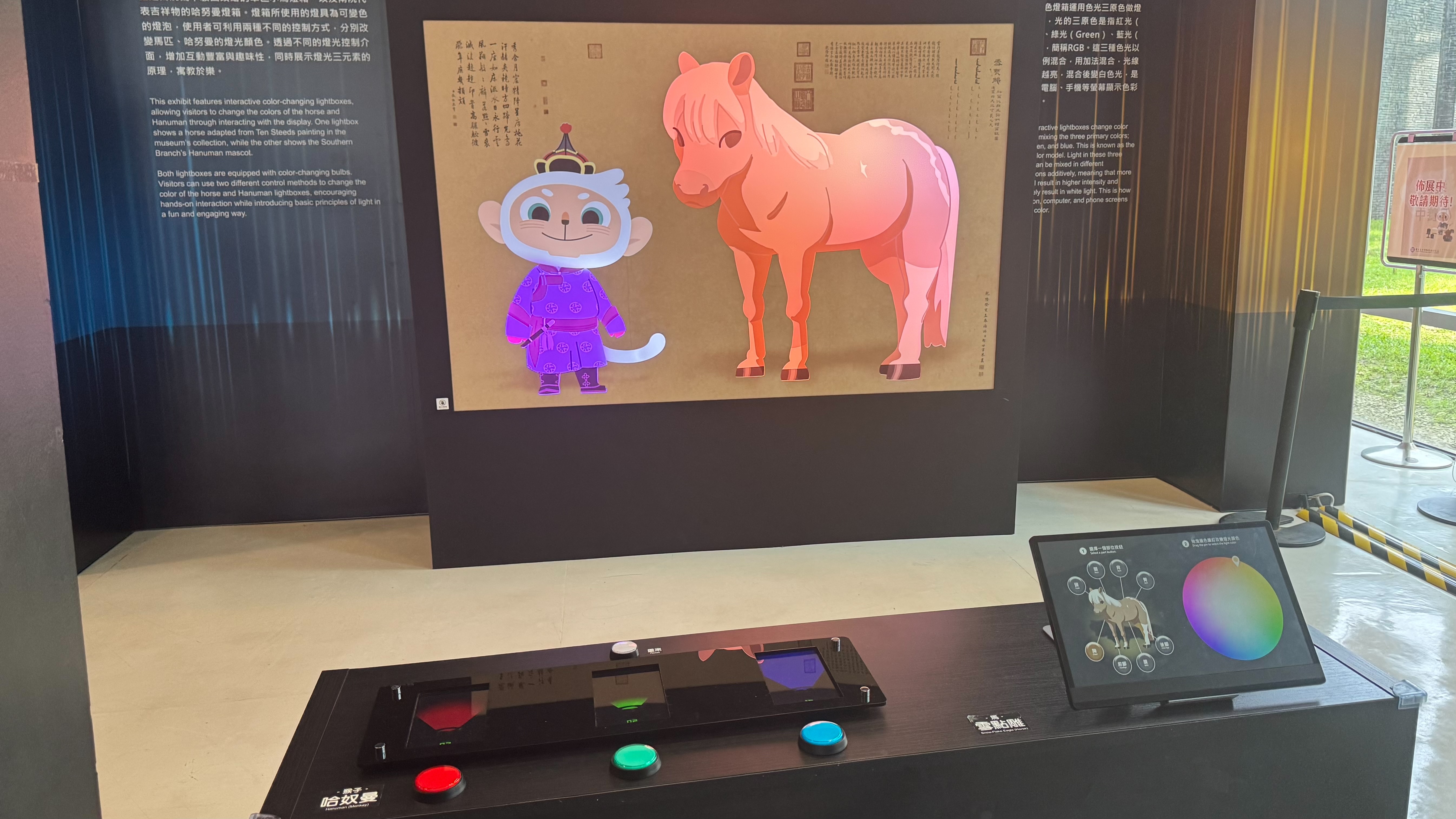 展區牆上有一幅Q版猴子與馬的背光畫作，前方設有紅、綠、藍三色按鈕的互動裝置與螢幕。