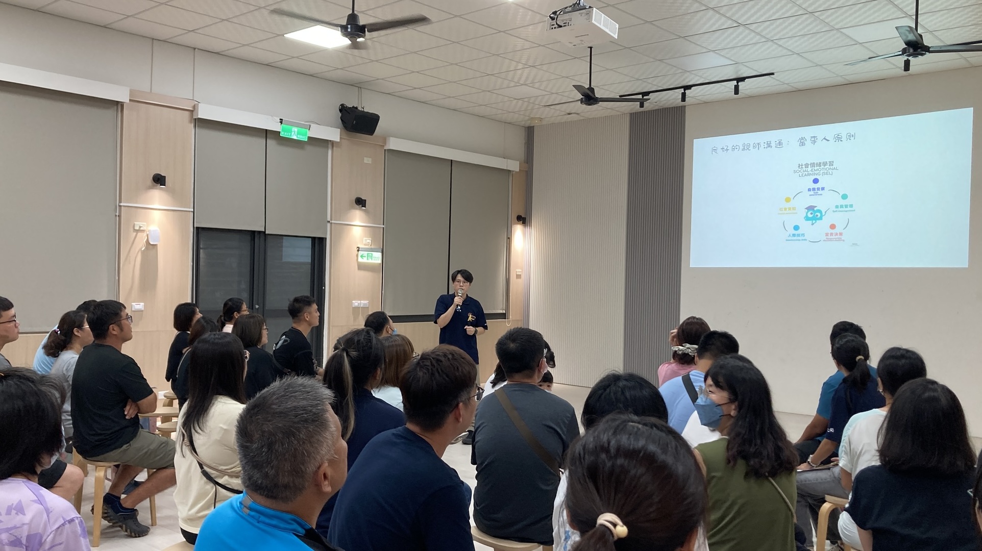 教室內坐滿學生家長，前方有一位老師站在投影牆前