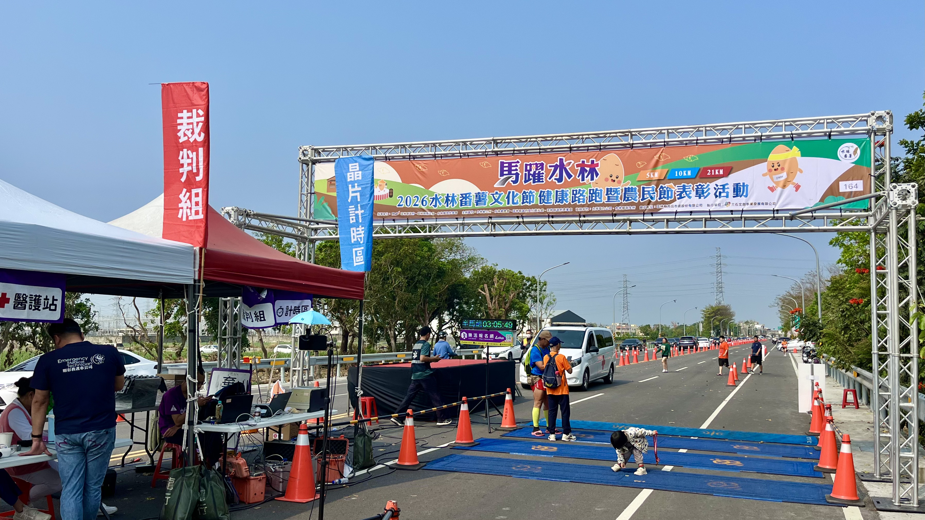 「馬躍水林」路跑活動現場實景，畫面包含活動拱門、裁判組帳篷、晶片計時區以及正在進行比賽的封閉道路與參賽選手。