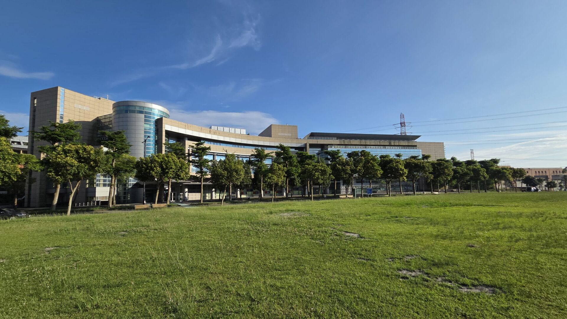 在晴朗的藍天下，一棟大型的現代化建築前方座落著一排綠樹與一大片寬廣的綠色草地。