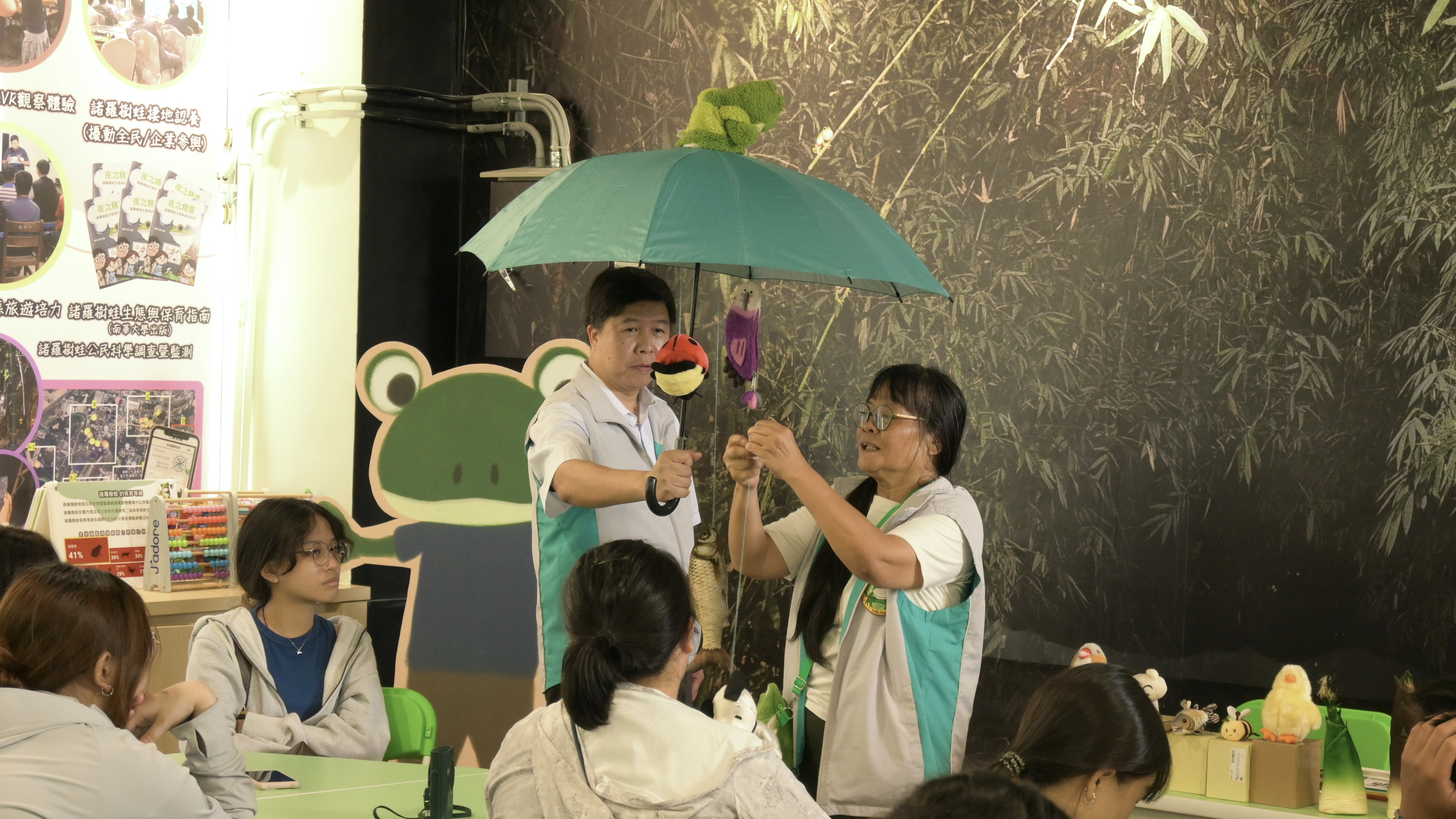 兩位解說員拿著雨傘和玩偶道具與台下的體驗者解說