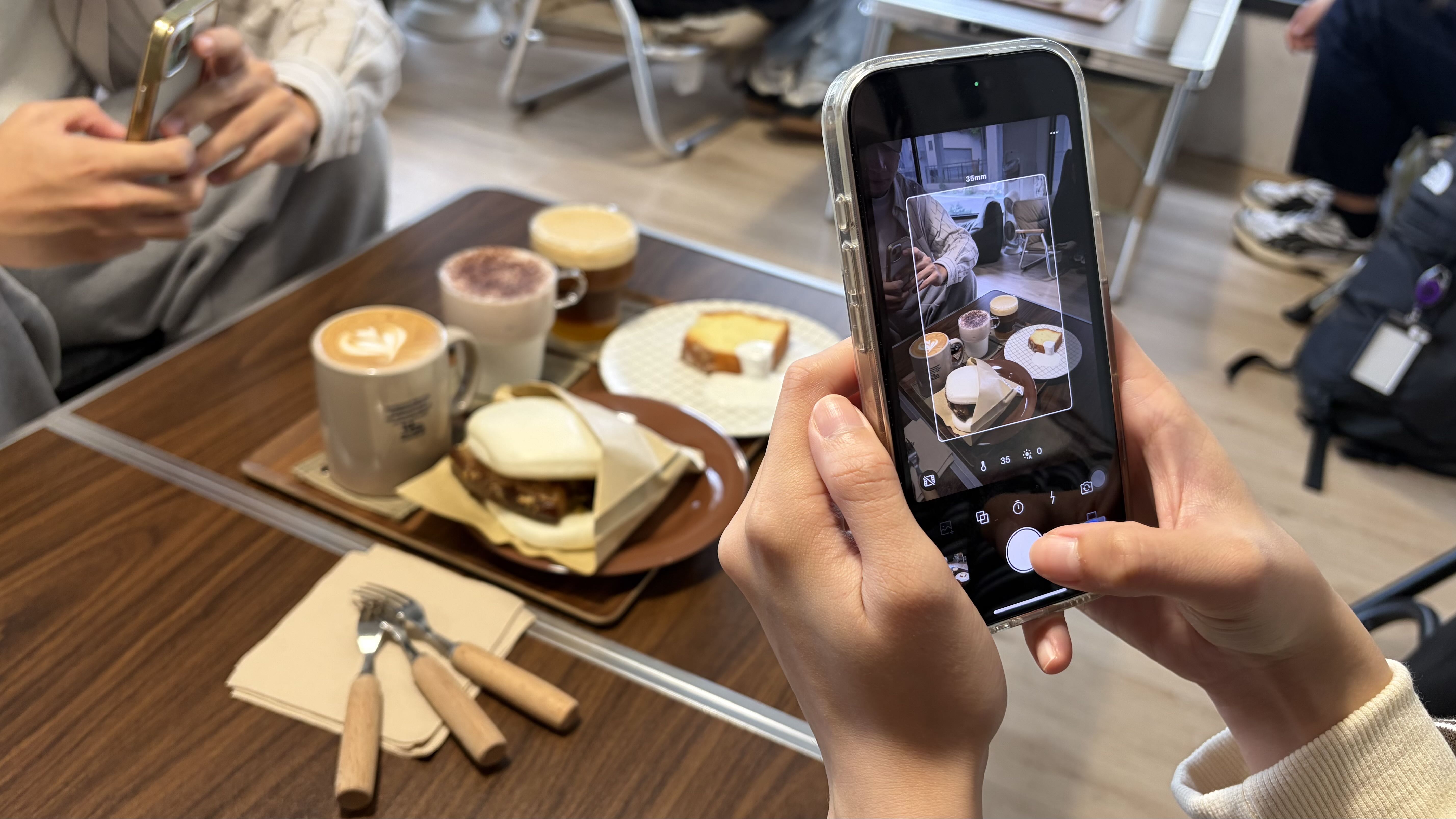 桌上擺放著飲料與餐點，兩個人拿著手機拍攝桌上的食物