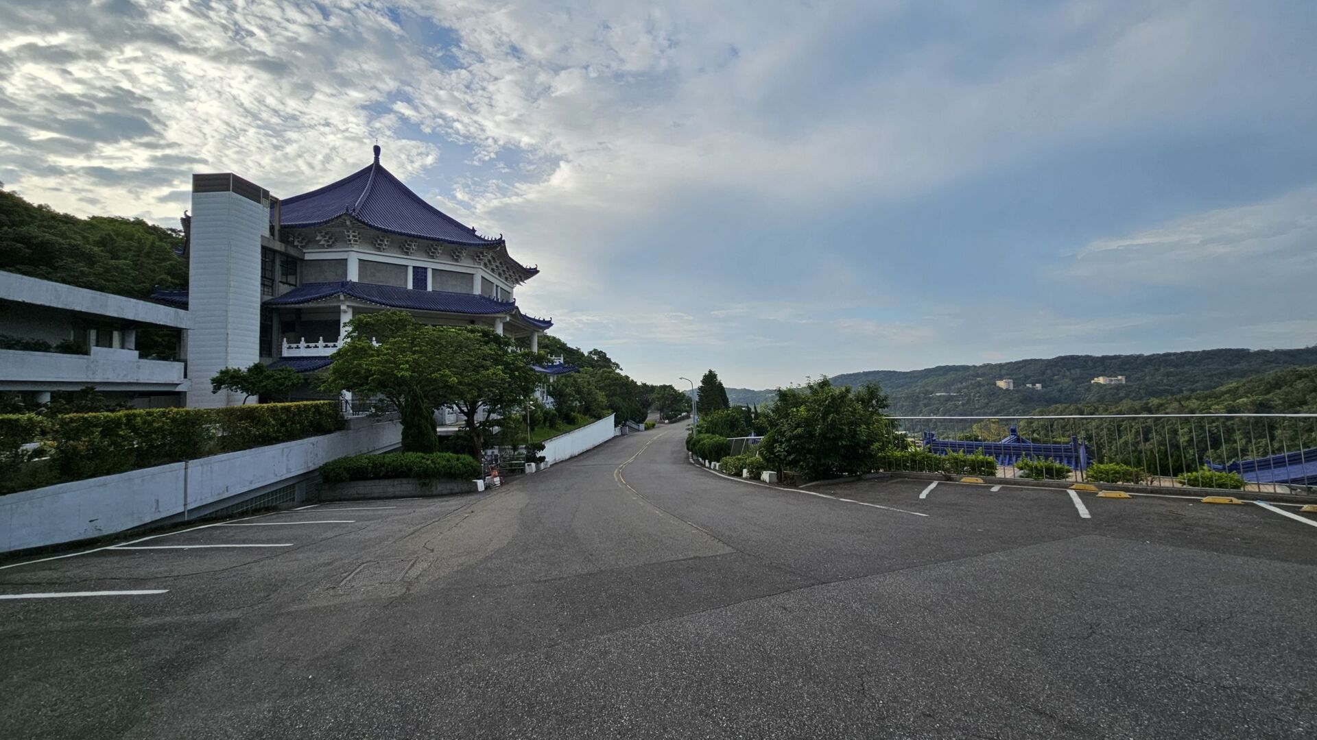 一棟擁有藍色傳統中式屋頂的大型建築座落於山林邊緣，前方為一條寬闊的柏油道路與停車場，遠處則可見連綿的山丘景致。