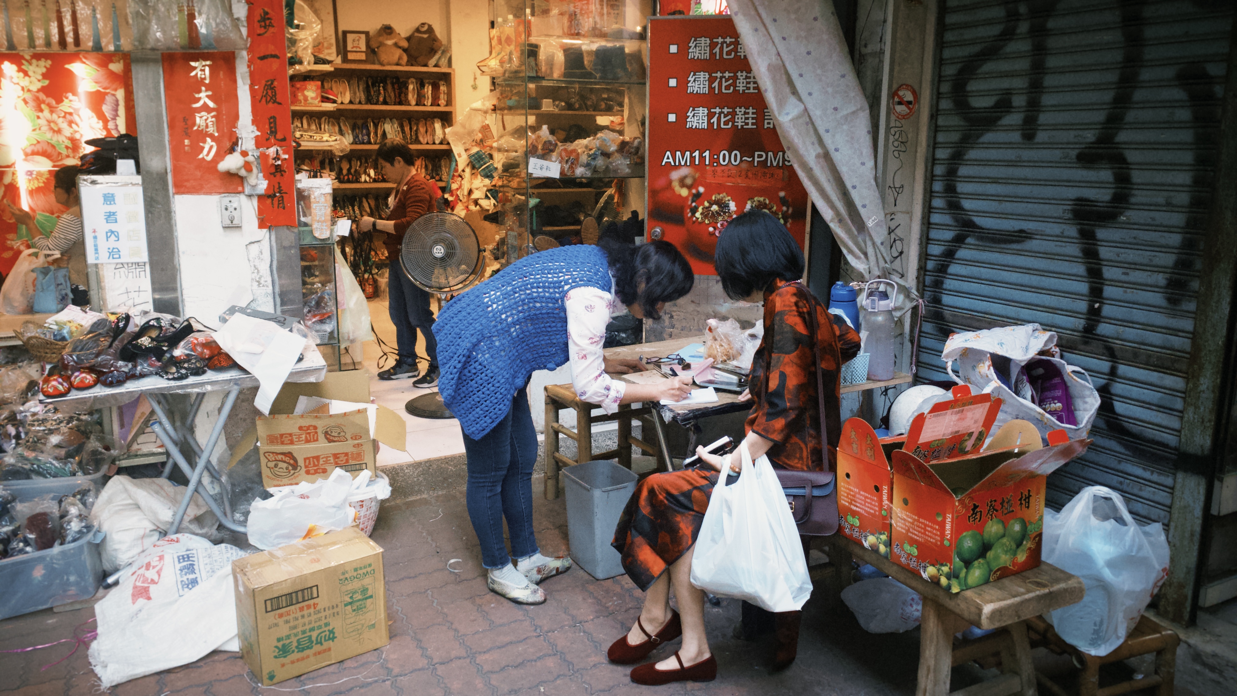 一名女子坐在店家門口的椅子上，另一名女子在一旁的桌子上寫字