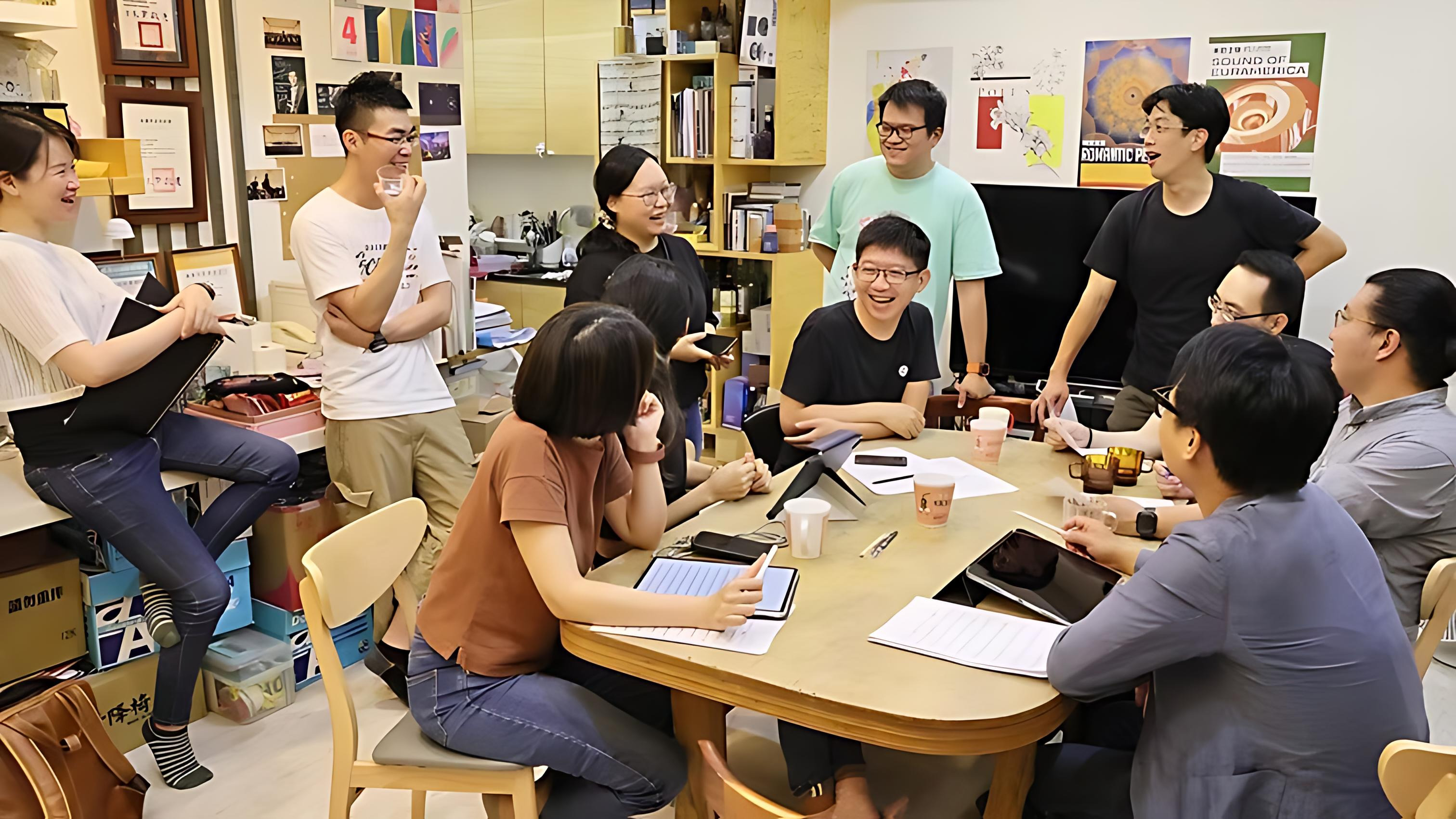 一群人聚集在貼滿藝文海報的工作室裡，圍繞著桌子輕鬆愉快地進行討論與交流。