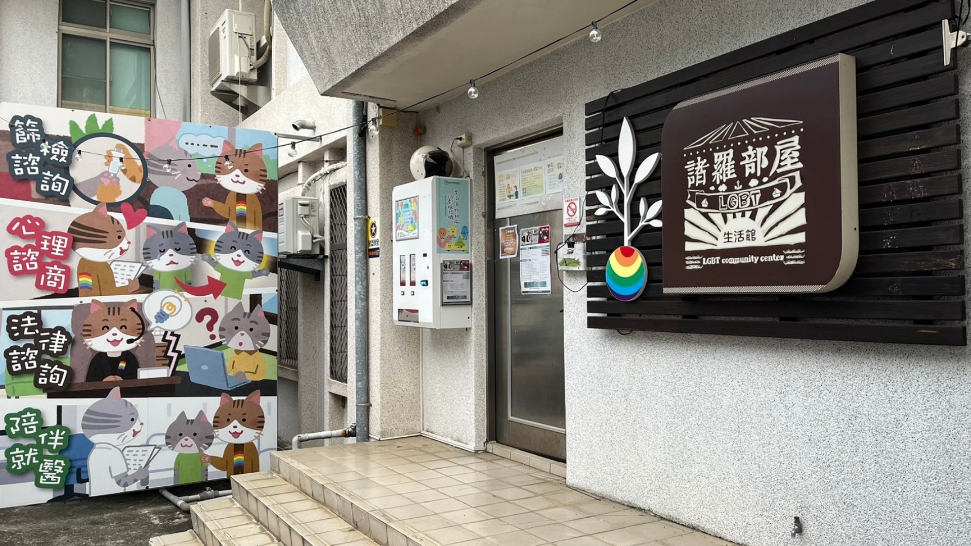 衛生所門口的右邊有諸羅部屋LGBT生活管的招牌，左方有篩檢諮詢、心理諮商、法律諮詢、陪伴就醫的字樣與插圖