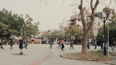 一個公園廣場裡許多學生跟著隊伍行走，一名學生拿著手機拍照